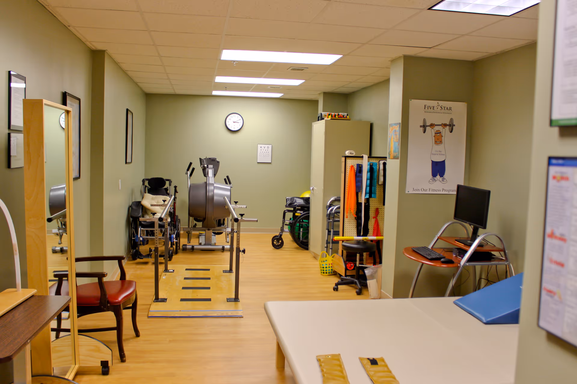 A rehabilitation or physical therapy room with exercise equipment, wheelchairs, parallel bars for walking practice, a treatment table, a computer workstation, and various therapy tools organized on a pegboard. The room has light green walls, a clock on the far wall, and a poster promoting a fitness program.