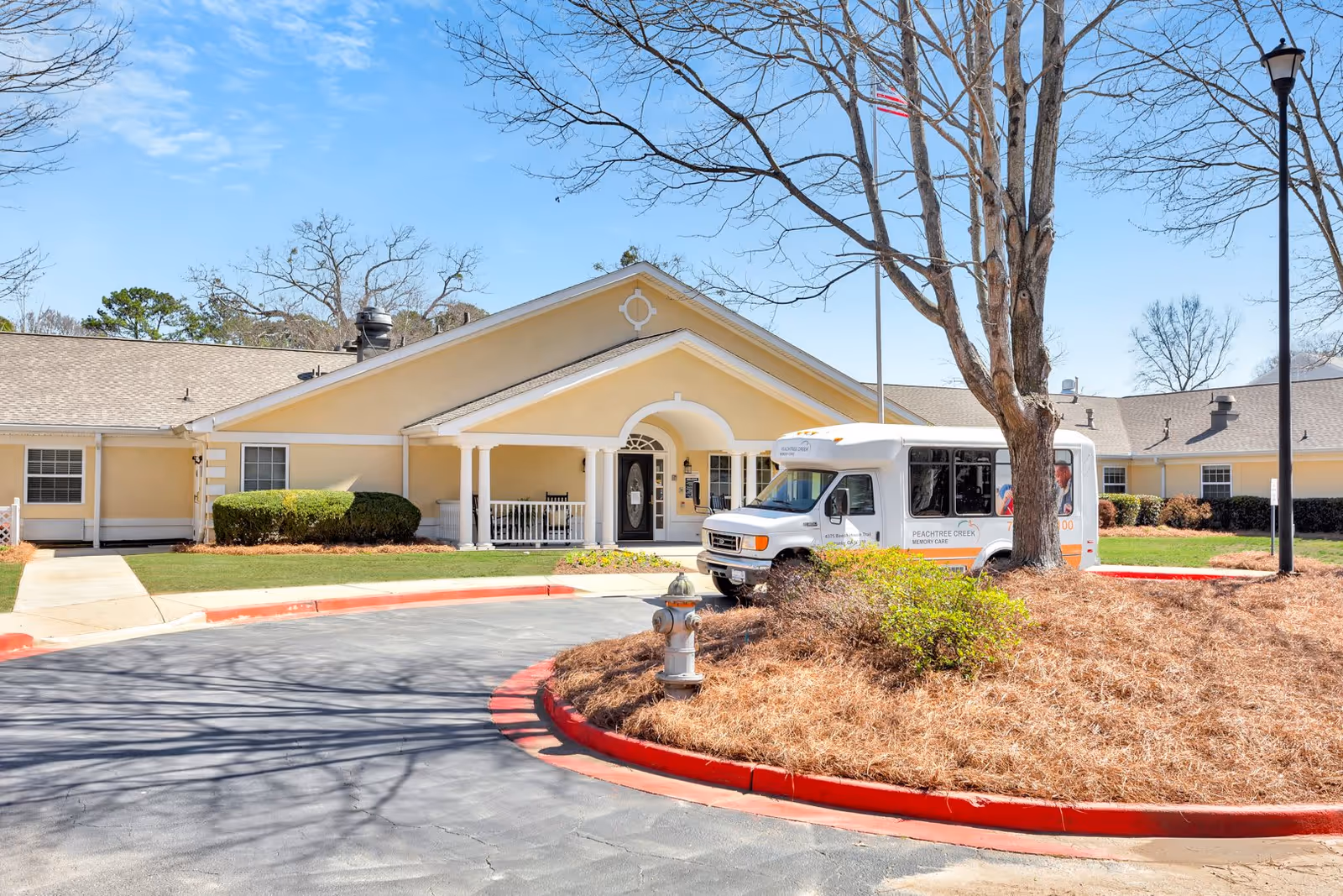 Exterior view of Peachtree Creek Memory Care assisted living facility in Georgia, showing a single-story beige building with a covered entrance, a white shuttle bus parked nearby, a flagpole with an American flag, and a landscaped roundabout with a fire hydrant and some bushes.