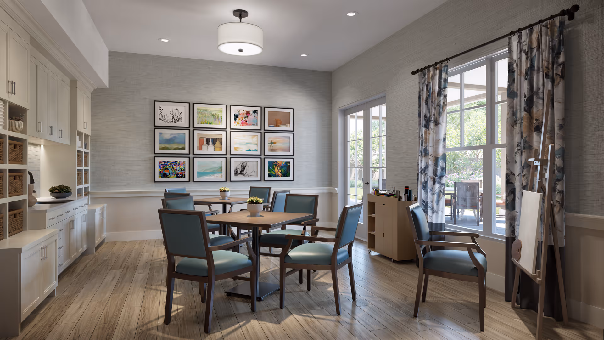 A bright and inviting common area with wooden flooring, a round table surrounded by six blue cushioned chairs, and a wall decorated with multiple framed artworks. There is a large window with floral curtains letting in natural light, and a small cabinet with art supplies next to an easel.