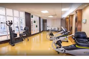 Interior view of a fitness room with exercise equipment including stationary bikes and elliptical machines. The room has large windows on one side allowing natural light to enter, yellow flooring, and white ceiling tiles with recessed lighting.