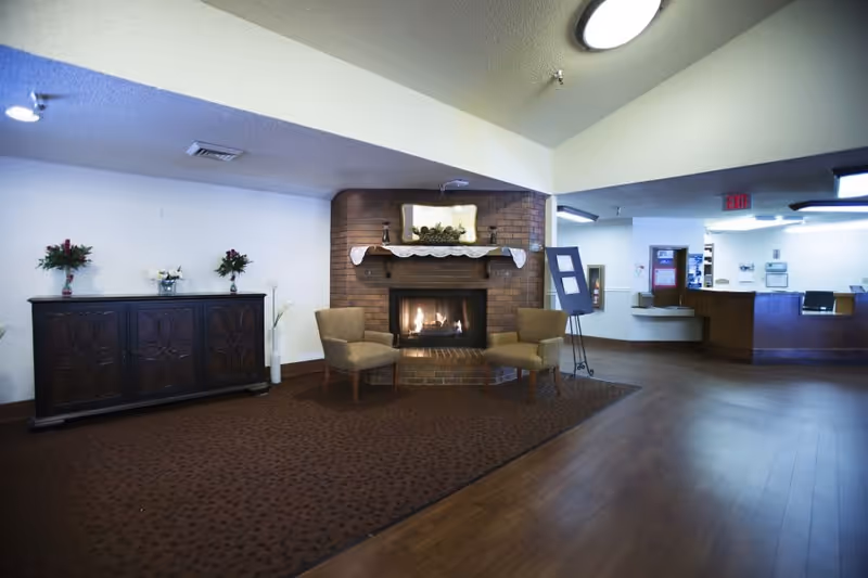 A cozy interior common area featuring a brick fireplace with a fire burning, two beige armchairs placed on a brown carpet in front of the fireplace, a dark wooden cabinet with flower arrangements on top against a white wall, and a reception desk area in the background.