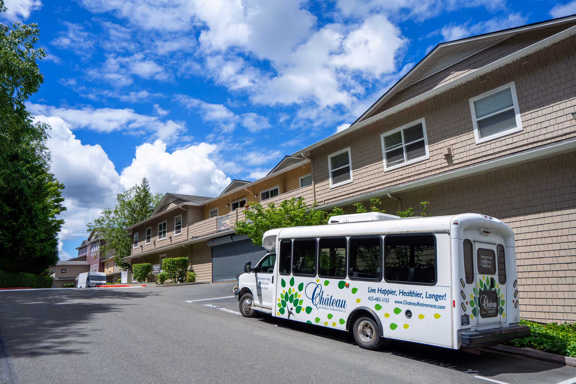 Exterior view of Chateau Bothell Landing Retirement facility on a sunny day with a partly cloudy sky. A white shuttle bus with the facility's name and slogan 'Live Happier, Healthier, Longer!' is parked on the driveway in front of the building. The building has beige siding and multiple windows, surrounded by trees and greenery.