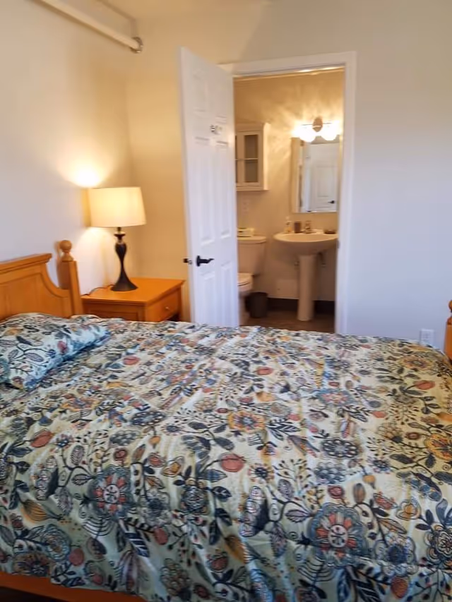A bedroom with a wooden bed frame and a floral patterned bedspread. Next to the bed is a wooden nightstand with a lamp. The door to an adjoining bathroom is open, showing a toilet, pedestal sink, mirror, and a wall-mounted cabinet.