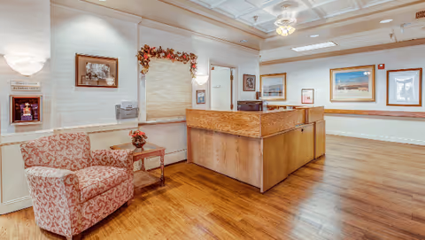 Reception area with a wooden front desk, upholstered armchair, side table and framed artwork in a well-lit interior.