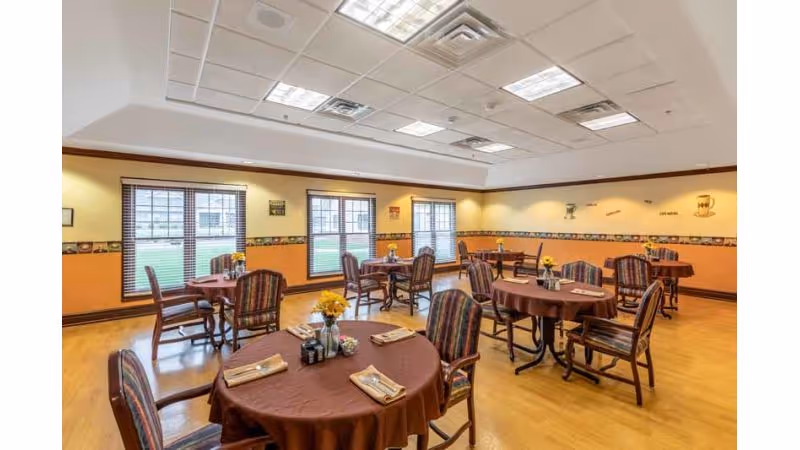 A dining room with several round tables covered with brown tablecloths, each set with napkins and small flower arrangements. The room has large windows with blinds, yellow and orange walls with decorative border trim, and wooden chairs with striped cushions.