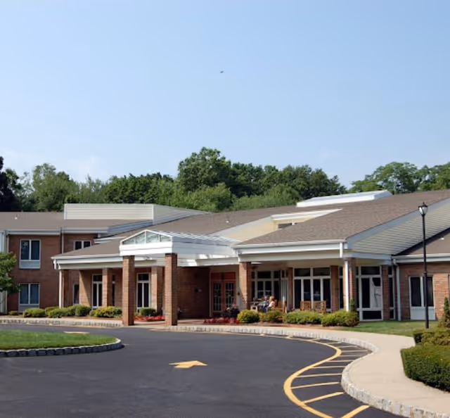 Front exterior view of a single-story assisted living facility building with brick walls, large windows, a covered entrance supported by brick columns, and a curved driveway with yellow road markings. There are some shrubs and greenery around the building and trees in the background under a clear blue sky.