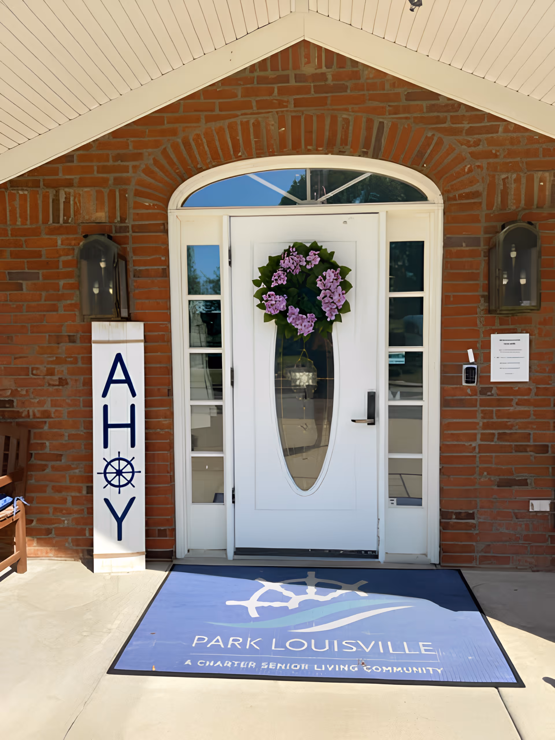 Entrance door of a senior living community with a white door adorned with a purple flower wreath. The door is set in a red brick wall with two lantern-style lights on either side. A vertical sign next to the door reads 'AHOY' with a ship wheel symbol replacing the letter O. A blue welcome mat on the ground in front of the door displays the text 'PARK LOUISVILLE A CHARTER SENIOR LIVING COMMUNITY' along with a ship wheel and wave graphic.
