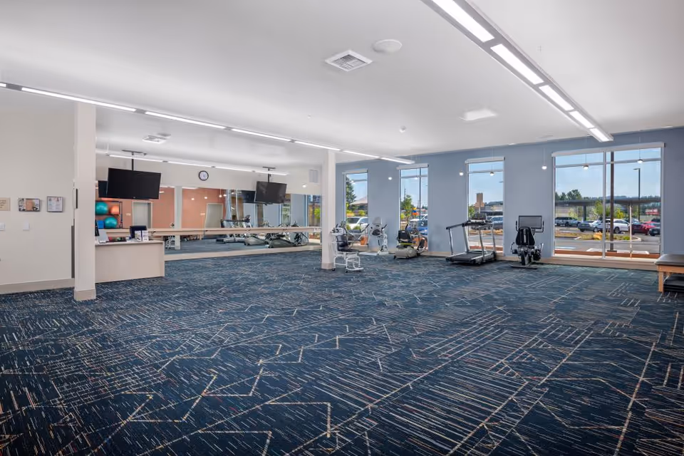 A spacious fitness room with patterned blue carpet, exercise equipment including a treadmill, stationary bikes, and weight machines. Large windows allow natural light to fill the room, and two flat-screen TVs are mounted on the wall above a counter. A large mirror spans one wall, reflecting the equipment and room interior.