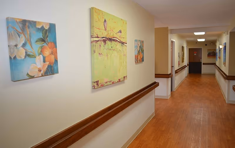 Well-lit decorated hallway in a senior living facility with wood handrails and framed artwork along the walls.