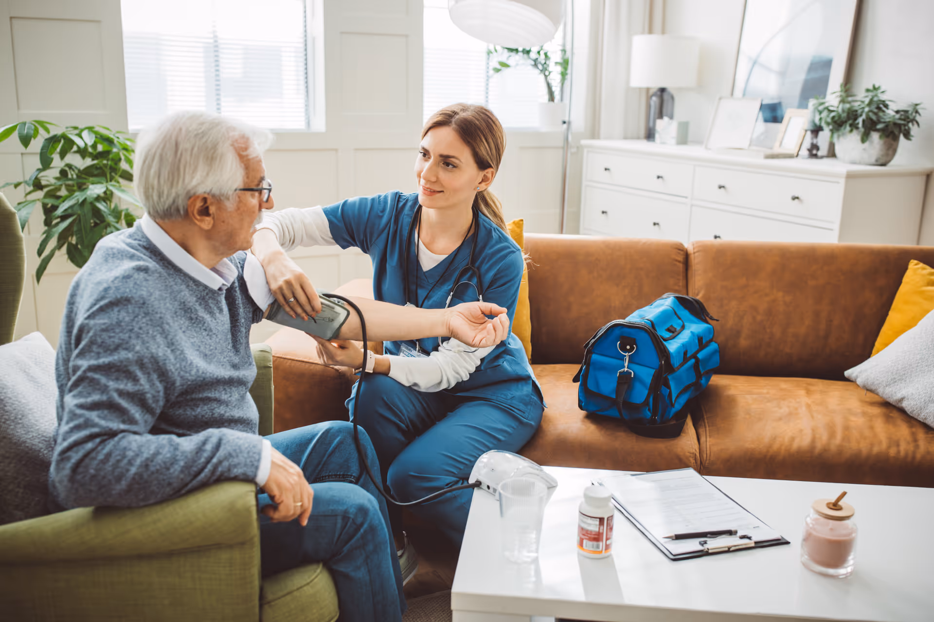 A healthcare professional in blue scrubs is measuring the blood pressure of an elderly man sitting on a green armchair in a well-lit living room. The room features a brown sofa, a white coffee table with medical supplies, and plants near the window.