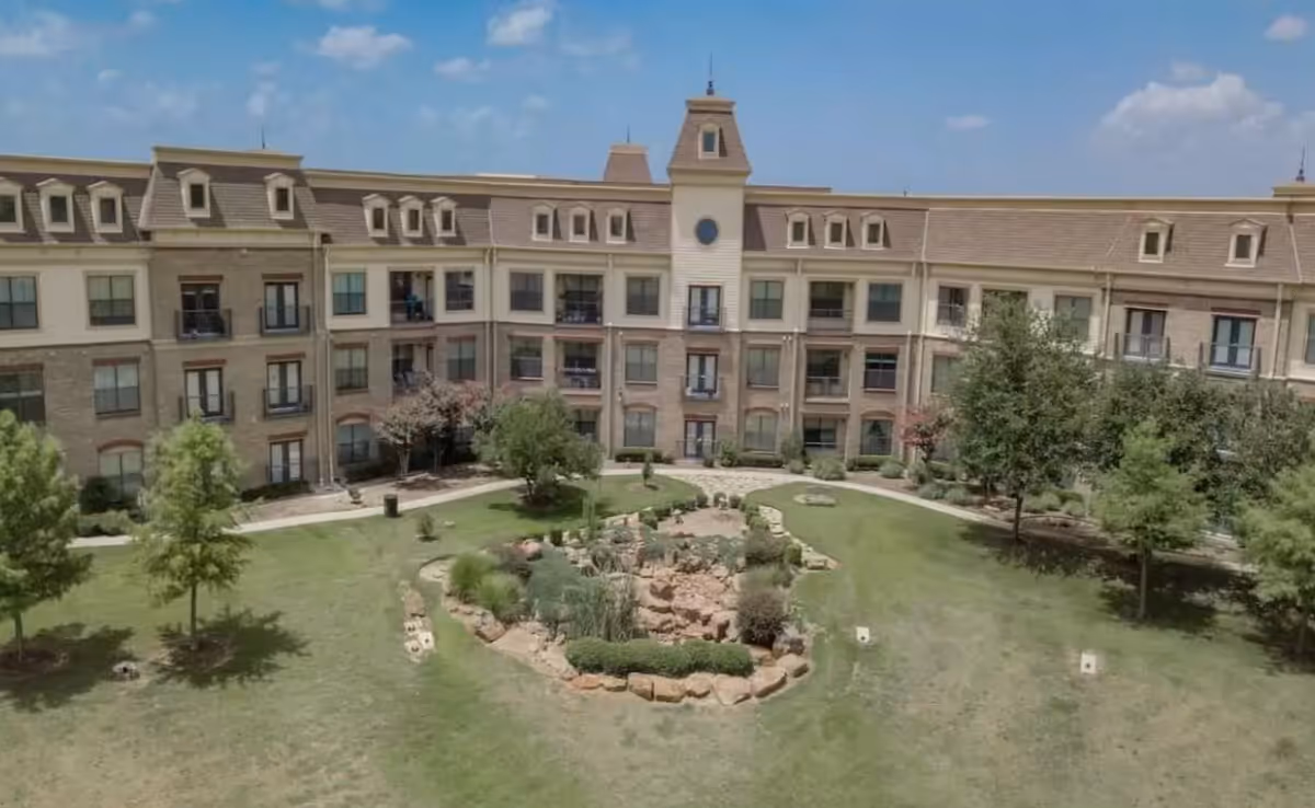 Front exterior of a multi-story senior living building overlooking a landscaped courtyard with lawn, trees, and a central rock garden.