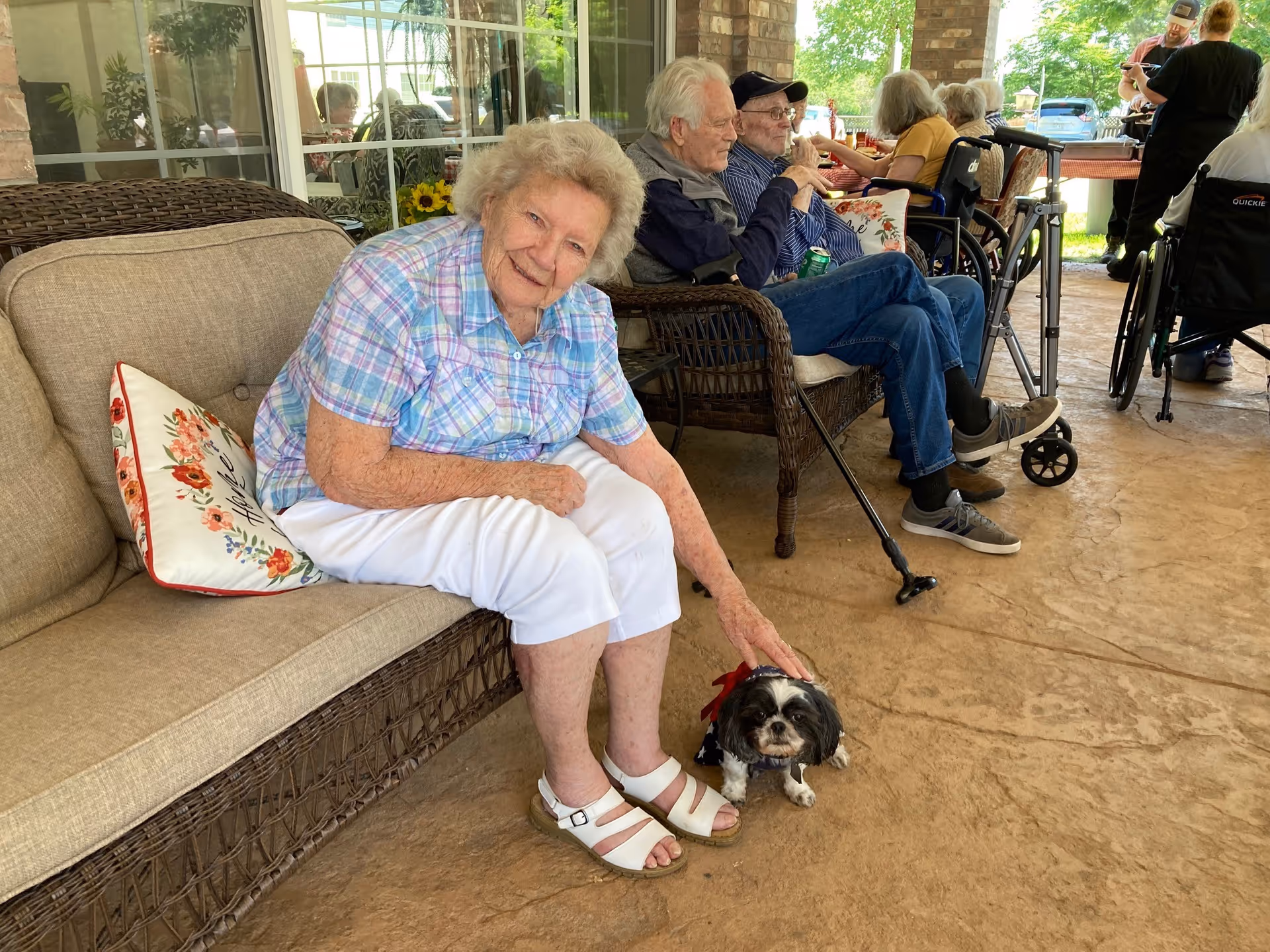 An elderly woman sitting on a cushioned wicker bench on a covered patio, smiling and petting a small black and white dog wearing a red, white, and blue outfit. Other elderly people are seated on similar wicker chairs in the background, some in wheelchairs, with a table and greenery visible outside.