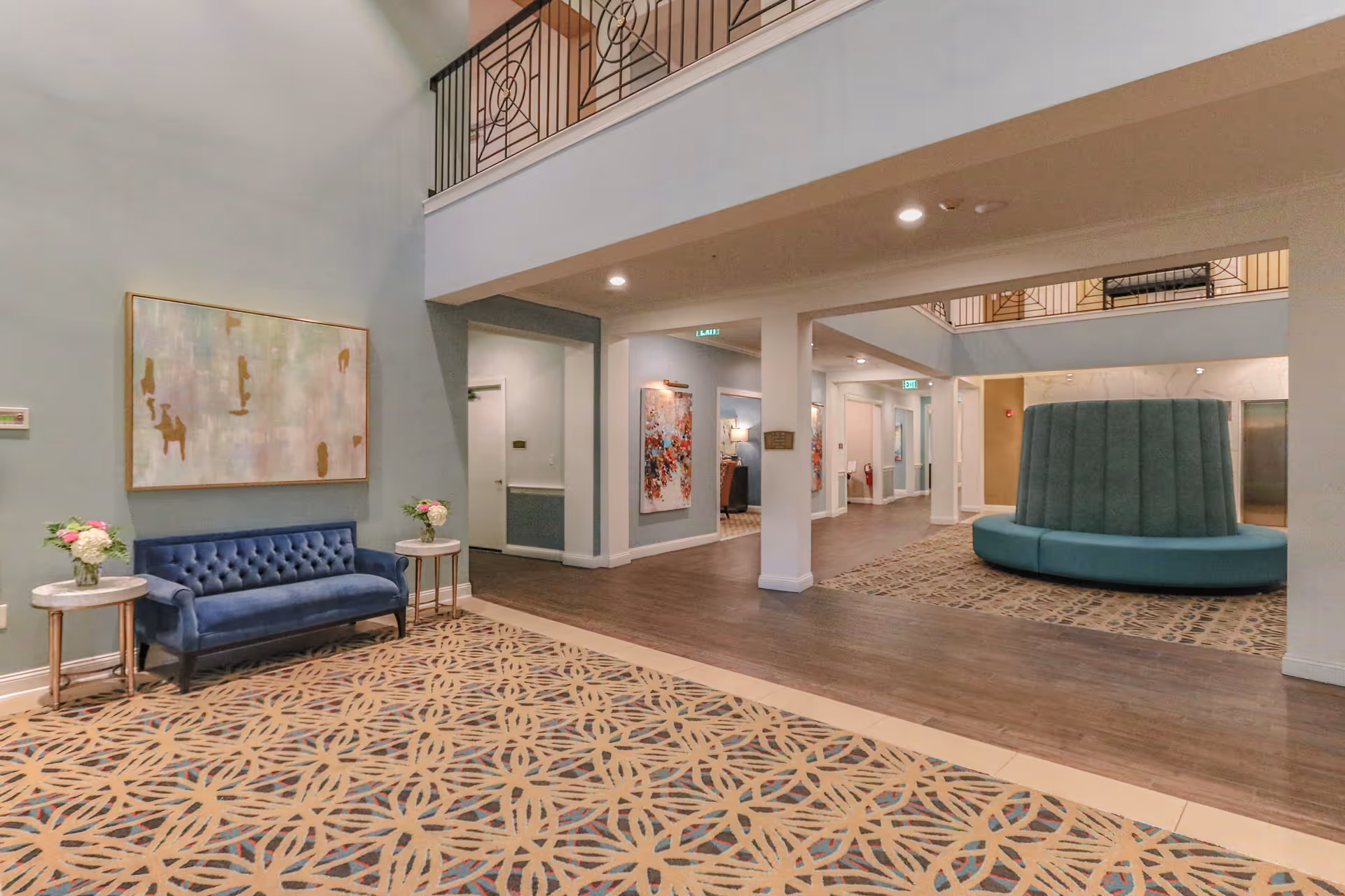 Interior view of a senior living facility lobby with a blue tufted sofa flanked by two small round tables holding flower arrangements. The floor has a patterned carpet, and there is a large circular teal seating area in the background near an elevator. The walls are decorated with abstract paintings, and the ceiling has recessed lighting with a second-floor balcony visible above.