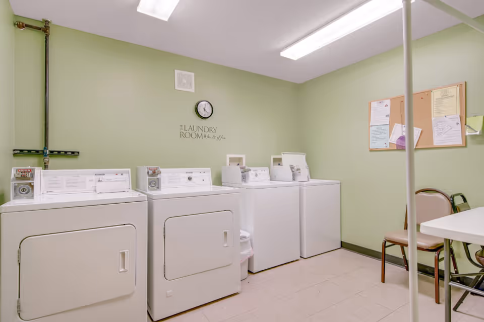 Laundry room with four white coin-operated washing machines and dryers against a light green wall. A clock and a sign that reads 'The Laundry Room - Made of this' are mounted on the wall above the machines. There is a bulletin board with papers pinned on the right wall, and a folding table with two chairs is partially visible.