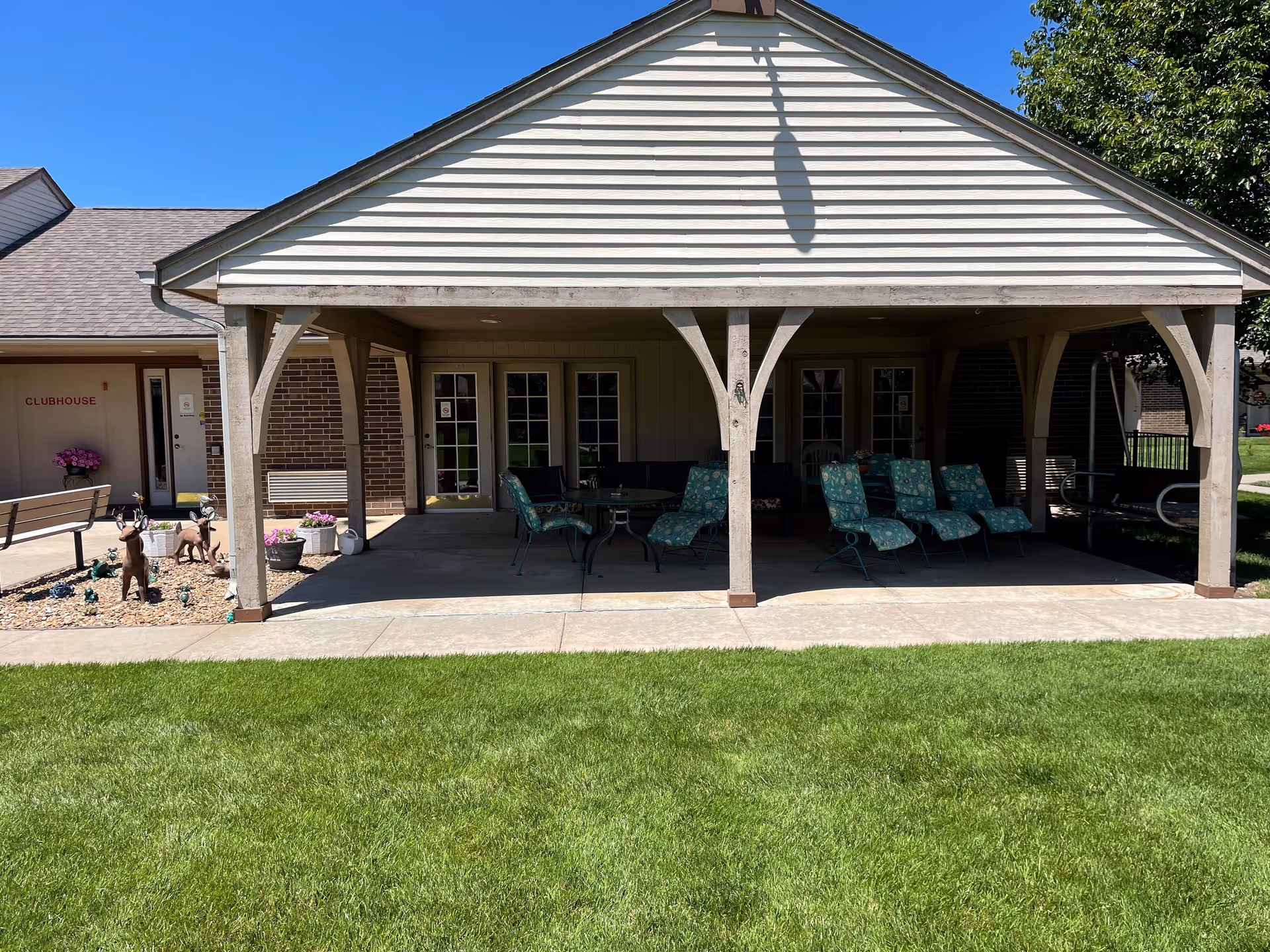 Covered outdoor patio area with several green cushioned chairs and a table, adjacent to a building with a sign that reads 'CLUBHOUSE'. The patio is supported by wooden beams and surrounded by a well-maintained lawn and some decorative plants and statues.