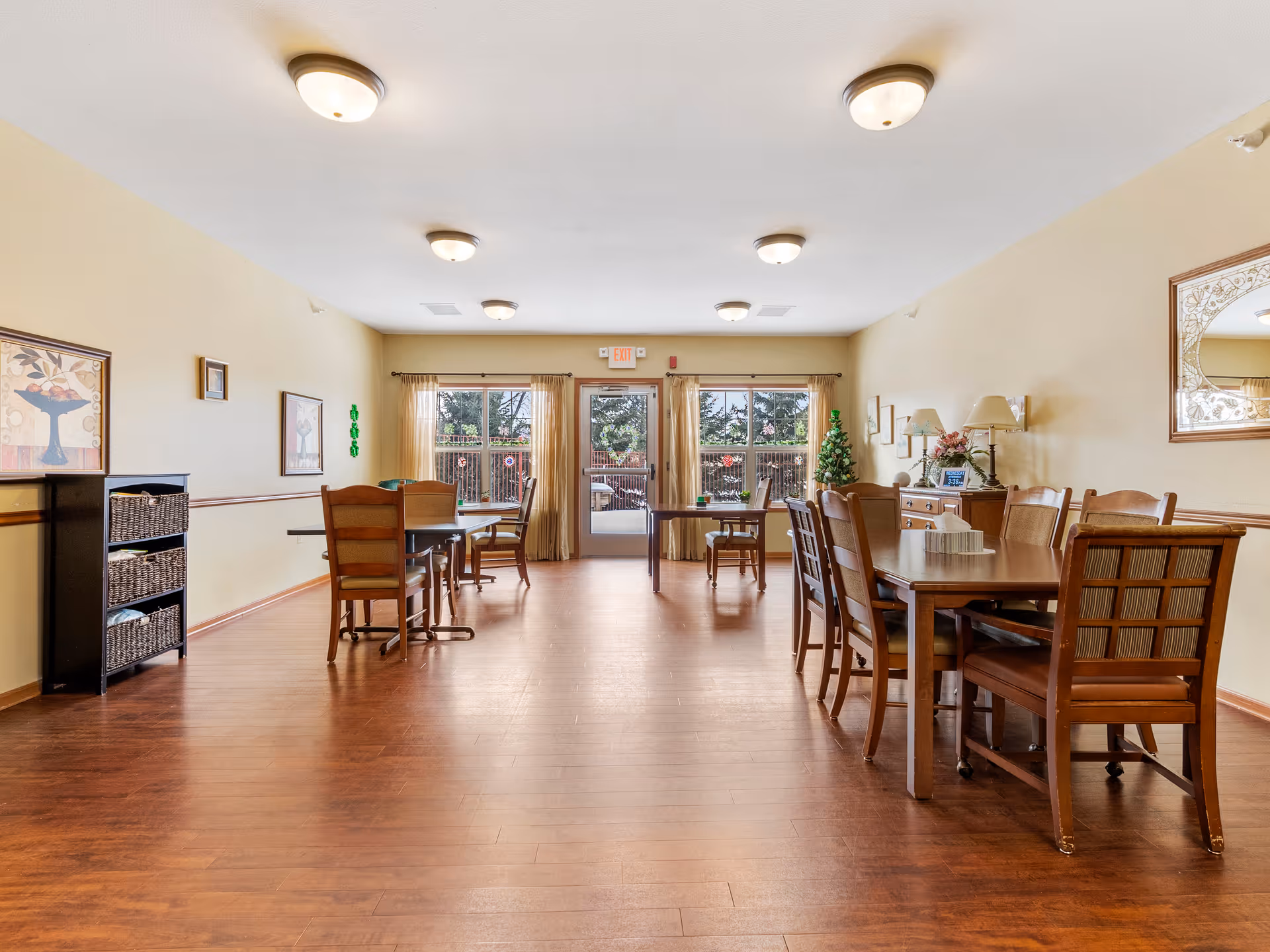 A spacious senior living dining room with wooden floors and multiple wooden tables and chairs. The room has beige walls adorned with framed artwork and a large mirror. There are ceiling lights evenly spaced, and large windows with sheer curtains letting in natural light. A small decorated Christmas tree and a sideboard with lamps and flowers are visible near the windows.