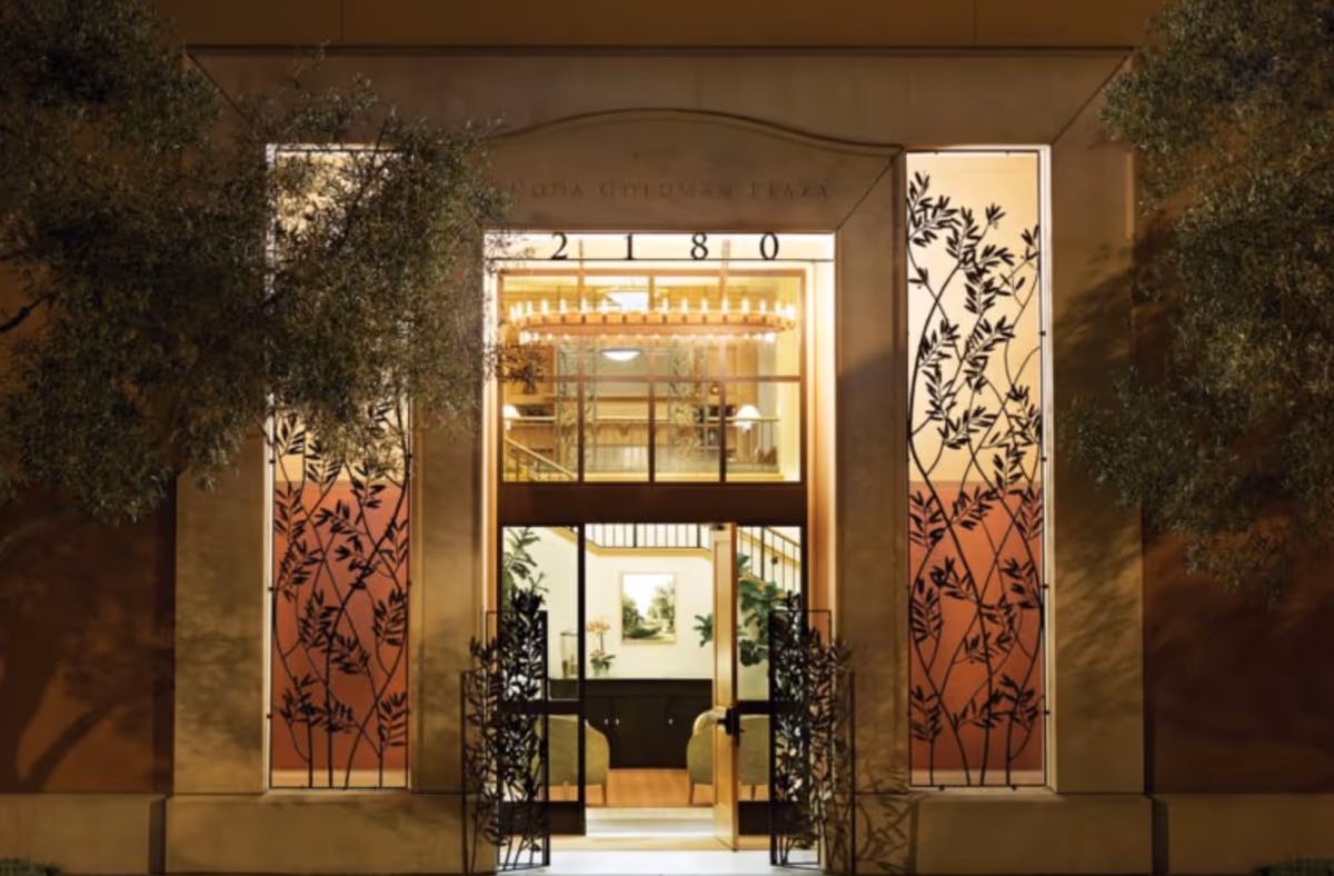 Night view of the entrance to Rhoda Goldman Plaza, a senior living facility, featuring an open glass door with decorative metalwork on either side, illuminated interior with plants, artwork, and a chandelier visible inside.