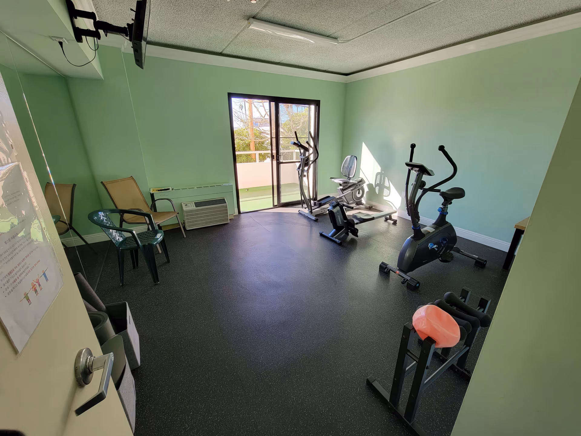 A small exercise room with light green walls and black rubber flooring. The room contains exercise equipment including a stationary bike, an elliptical machine, a rowing machine, and a recumbent bike. There are three chairs against the left wall and a glass door leading to an outdoor balcony. A wall-mounted TV is visible in the upper left corner.