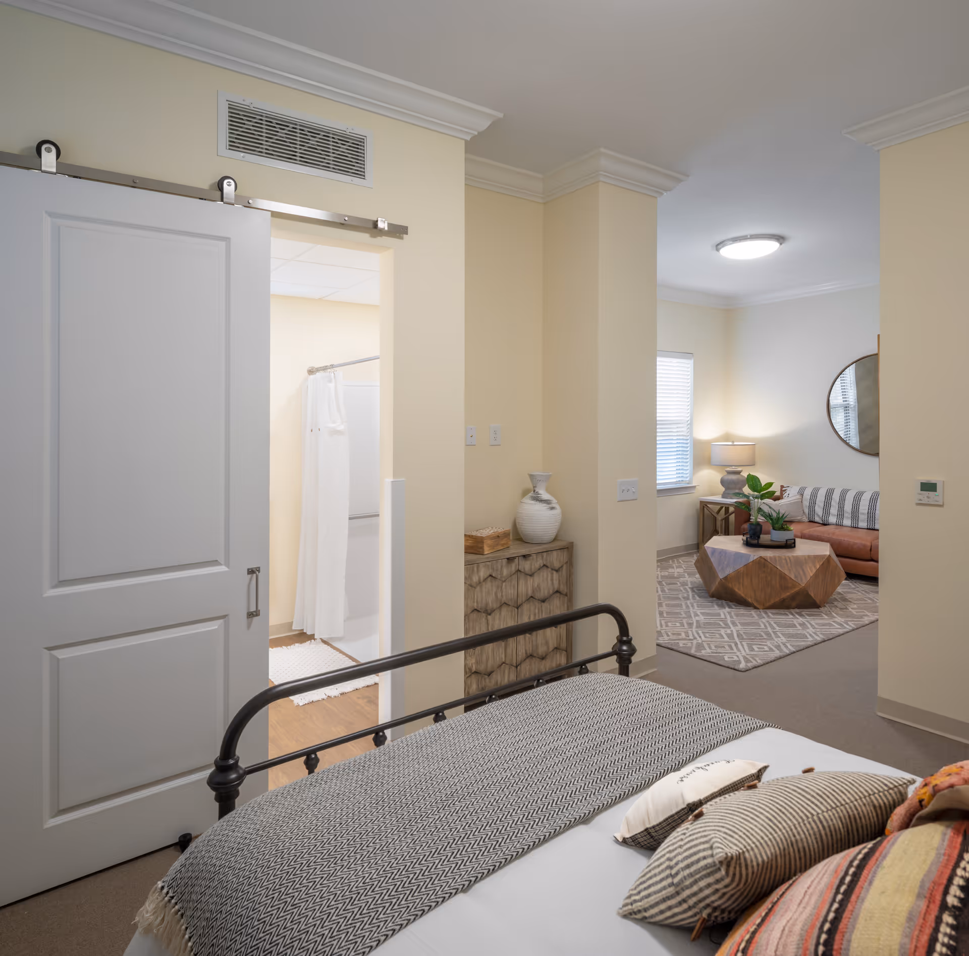 View from a bedroom showing a bed with patterned pillows and blanket, a sliding door leading to a bathroom with a shower curtain, and an adjacent living area with a sofa, round mirror, coffee table, and a lamp on a side table.