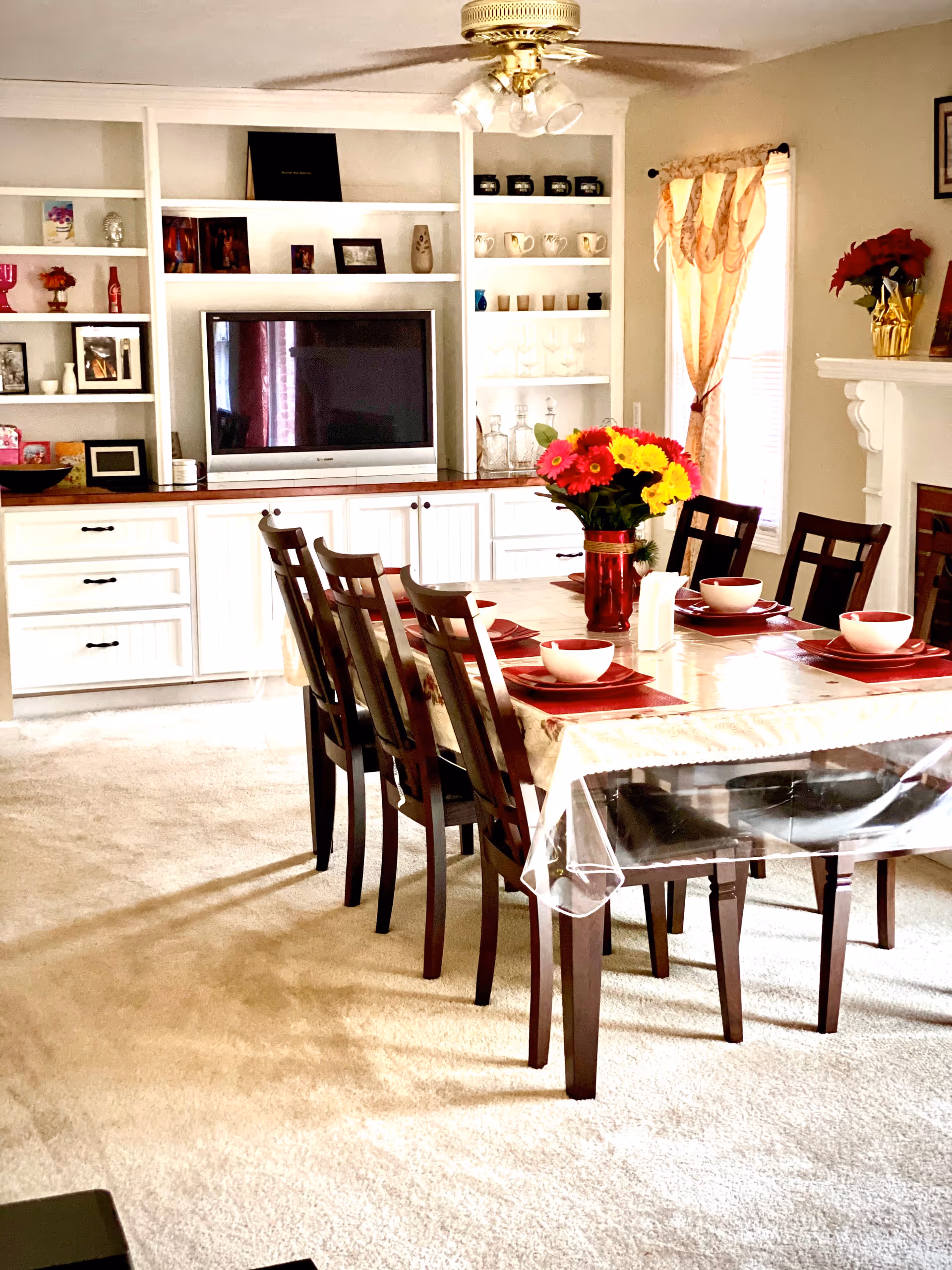 A dining room with a rectangular table covered with a tablecloth and a clear plastic cover. The table is set with red placemats, white bowls, and red plates. A vase with colorful flowers is in the center of the table. There are eight dark wooden chairs around the table. In the background, there is a built-in white shelving unit with various decorative items and a flat-screen TV. A window with sheer curtains and a fireplace with a flower pot on the mantel are also visible.