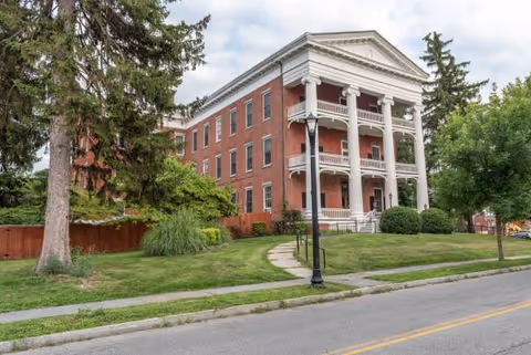 Brick, multi-story building with a white-columned front porch, surrounded by green lawn and trees.