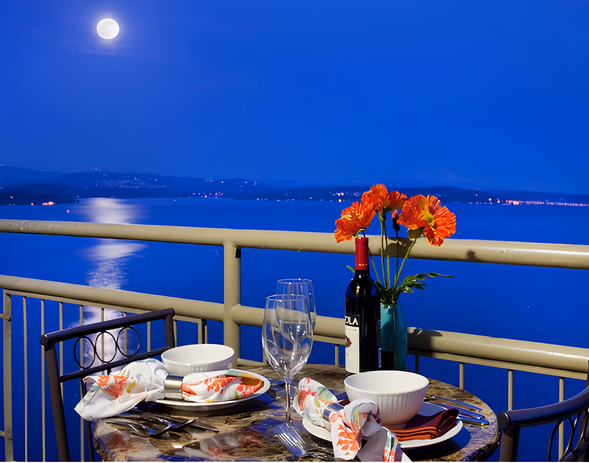 A small round table set for two on a balcony at night with a full moon reflecting on a large body of water. The table has two place settings with bowls, plates, floral napkins, silverware, two empty wine glasses, a bottle of red wine, and a vase with orange flowers. The balcony railing overlooks the water and distant hills with scattered lights.