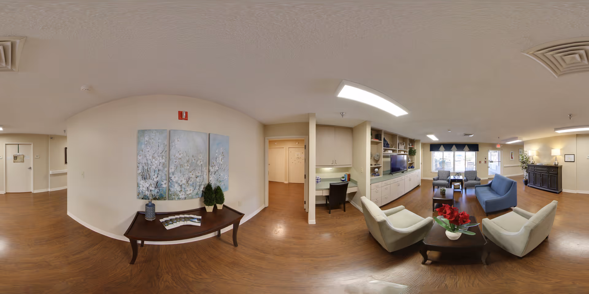 A spacious senior living facility common area with wooden flooring, beige walls, and ceiling lights. The room features a seating area with four armchairs arranged around a dark wooden coffee table with a vase of red flowers. There is a built-in cabinet with a TV and decorative items on shelves. A small desk with a chair is positioned near the cabinet. On the left side, a wooden table holds two small potted plants and some brochures, beneath a three-panel floral painting. Large windows with blue valances allow natural light into the room.