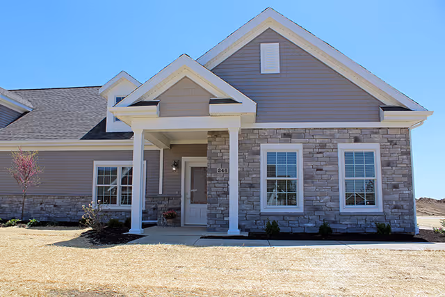Front exterior view of a single-story residential building with gray siding and stone accents, featuring a covered entrance with white columns and a door numbered 246, under a clear blue sky.