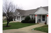 Exterior view of a single-story residential building with white siding and a gray roof. The building has a covered porch with a chair and potted plants. There is a concrete walkway leading to the entrance, green grass, and a leafless tree in the yard.