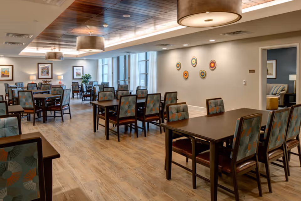 A spacious dining room with multiple dark wood tables and chairs featuring patterned upholstery. The room has a wooden floor and a ceiling with recessed lighting and large circular hanging light fixtures. The walls are decorated with framed artwork and colorful circular wall decorations. Large windows allow natural light to enter the room.