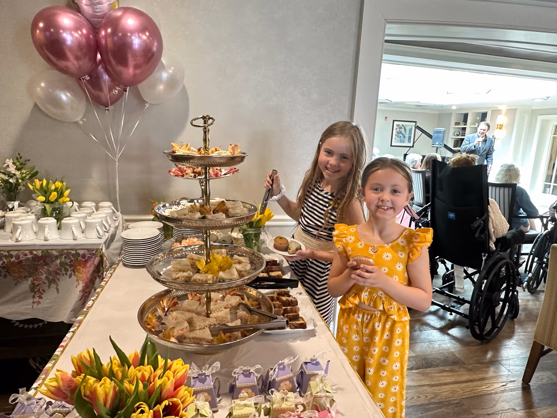 Two young girls stand at a decorated buffet table with tiered serving platters of sandwiches and desserts while seniors in wheelchairs are in the dining area behind them.