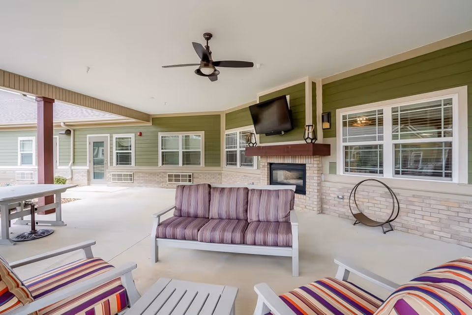 Covered outdoor patio area with cushioned seating including a sofa and chairs with striped upholstery, a ceiling fan, a wall-mounted TV above a fireplace, and green siding with brick accents on the walls.