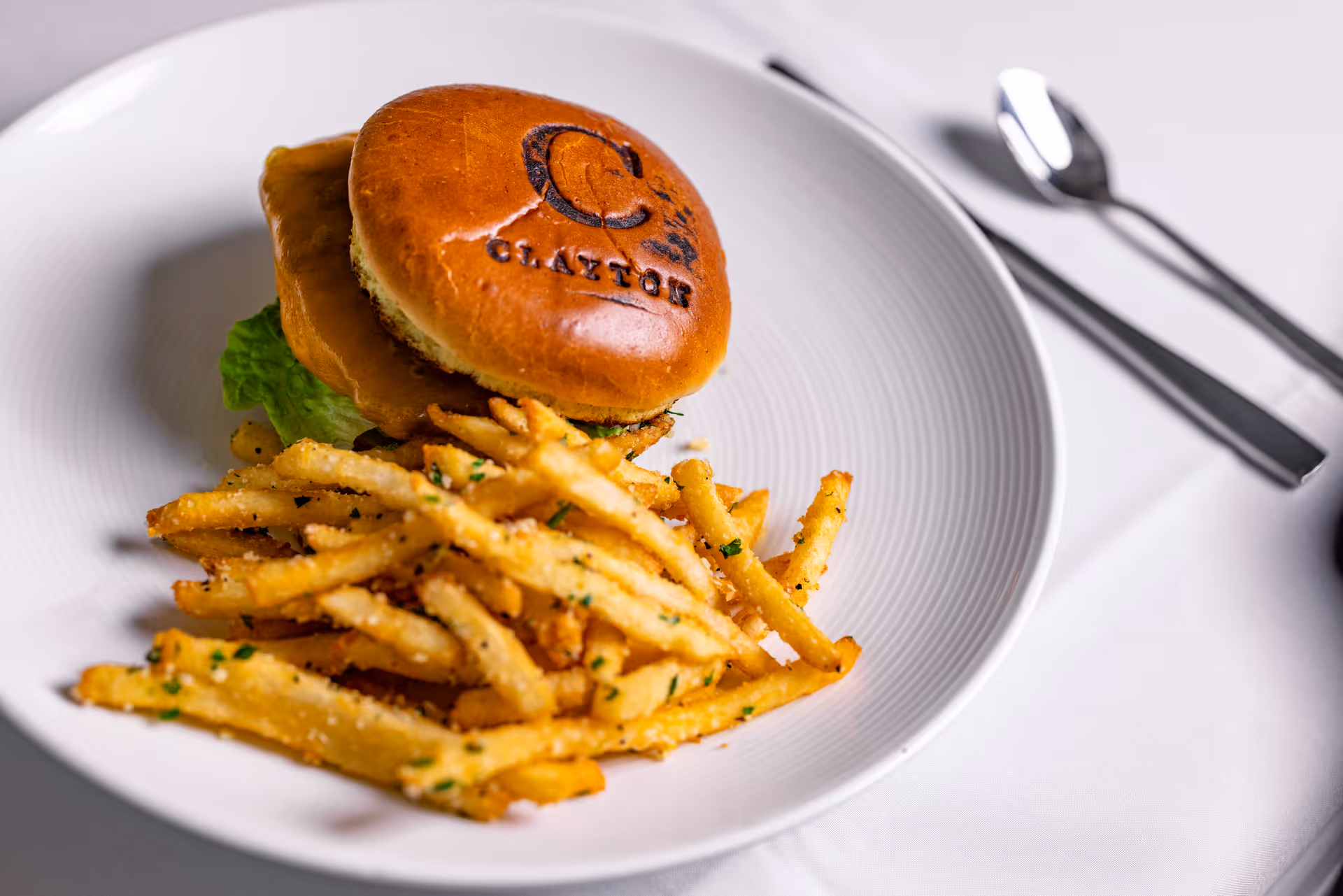 A plate with a sandwich featuring a bun branded with the Clarendale Clayton logo, served with a side of seasoned French fries on a white tablecloth with a fork and spoon in the background.