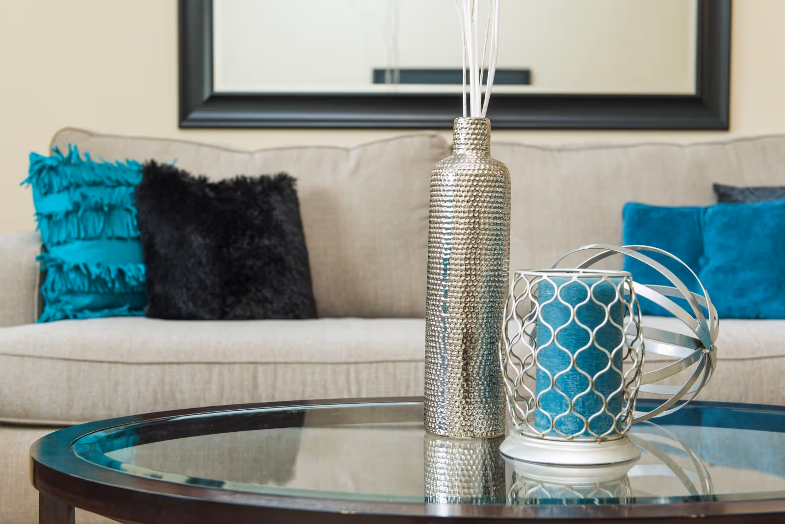 A beige sofa with decorative teal and black pillows in the background, and a glass coffee table in the foreground holding a textured silver vase, a blue candle in a silver holder, and a silver decorative orb.