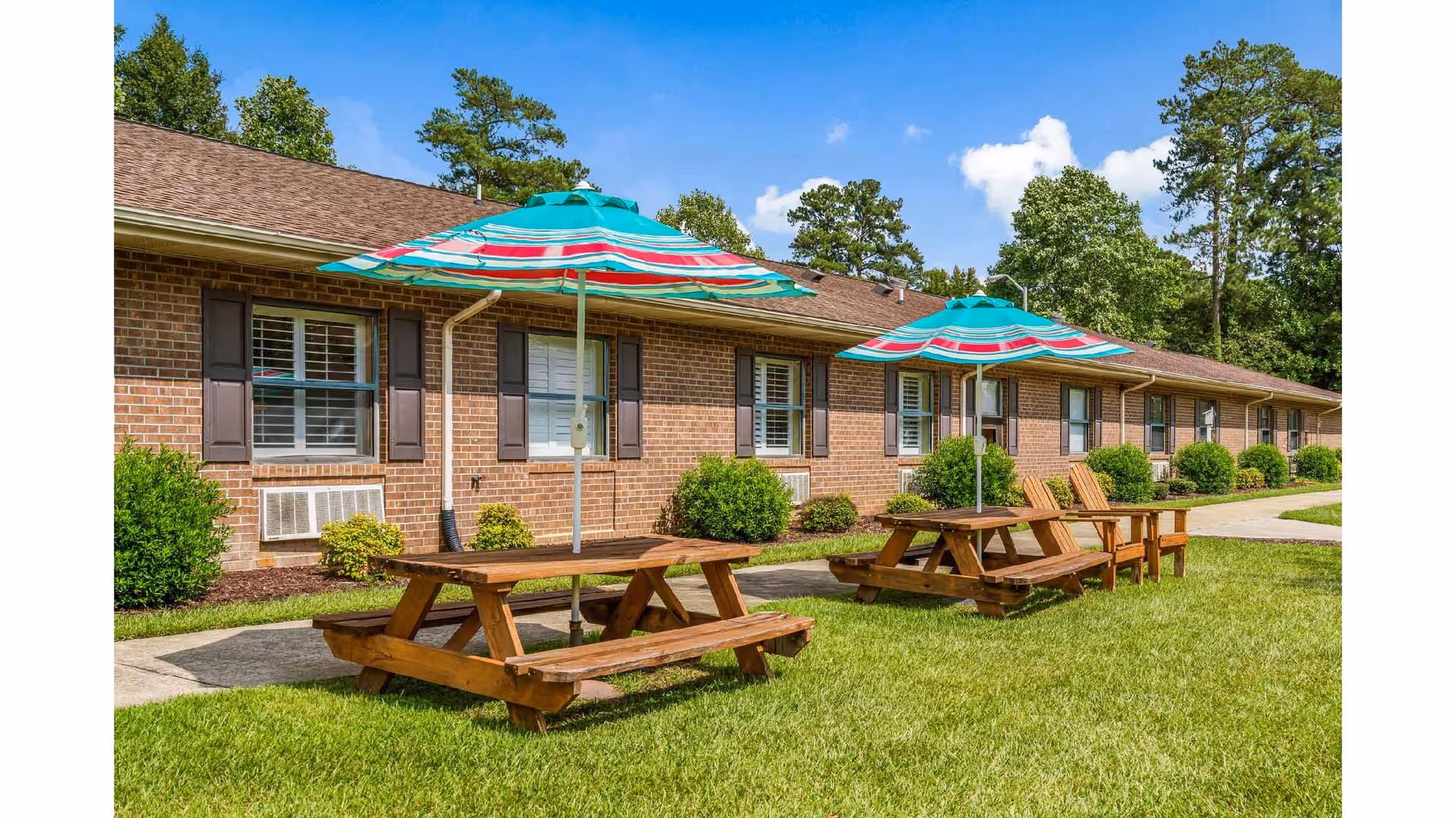 Outdoor area of Goldsboro Rehabilitation and Healthcare Center featuring two wooden picnic tables with colorful striped umbrellas on a grassy lawn in front of a brick building with windows and shrubs.