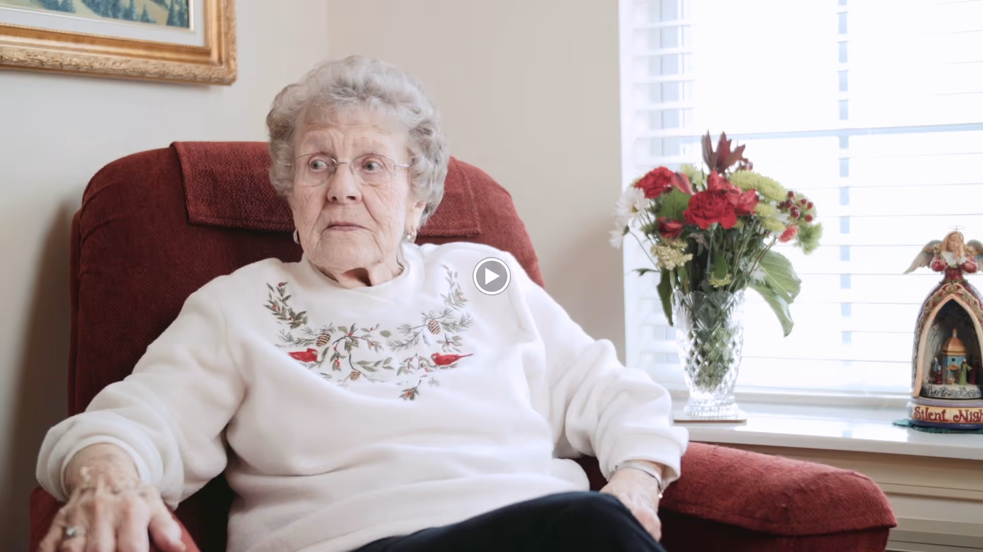 An elderly woman sits in a red armchair by a window with a vase of flowers on the windowsill.