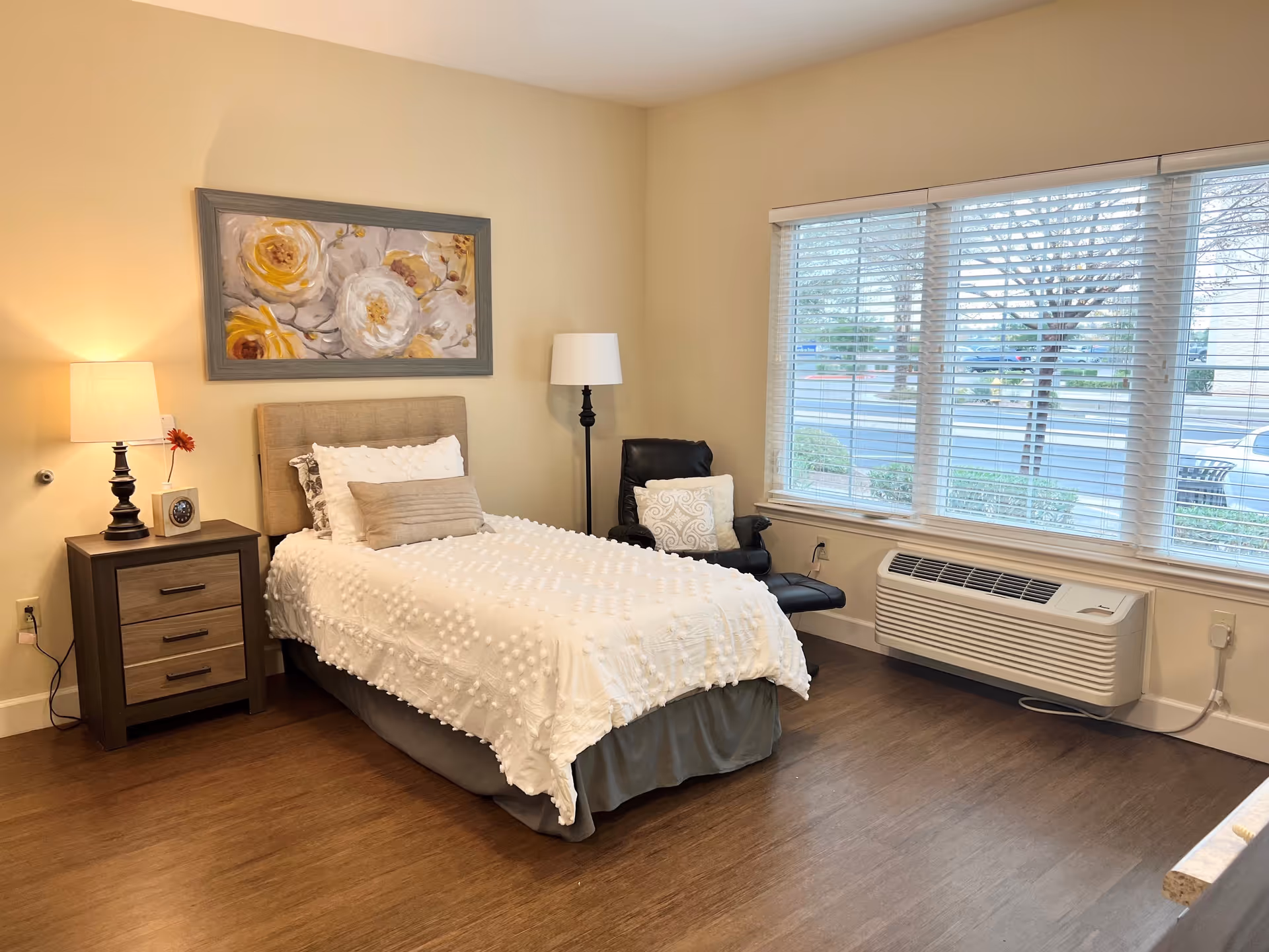 A cozy bedroom with a single bed covered in a white textured blanket and multiple pillows. Next to the bed is a wooden nightstand with a lamp, a clock, and a small flower. A framed floral painting hangs above the bed. There is a black armchair with a decorative pillow near a large window with white blinds. Below the window is a wall-mounted air conditioning unit. The room has wooden flooring and beige walls.