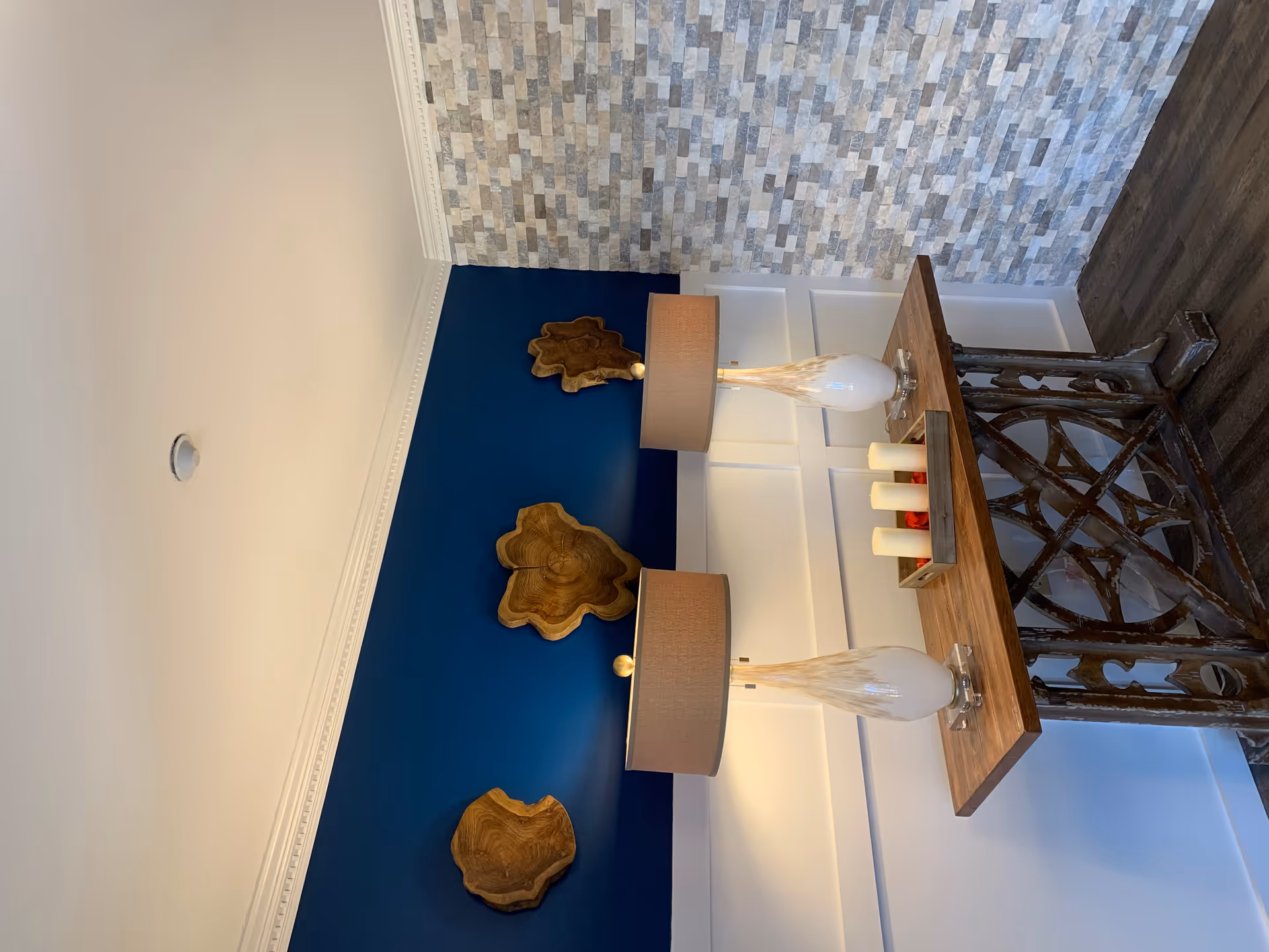 A decorative interior corner featuring a wooden console table with an intricate base, two matching table lamps with beige shades, and a tray holding four white candles. The wall behind the table is painted dark blue with three wooden art pieces mounted on it, and the adjacent wall is covered with textured gray stone tiles. The floor is dark wood.