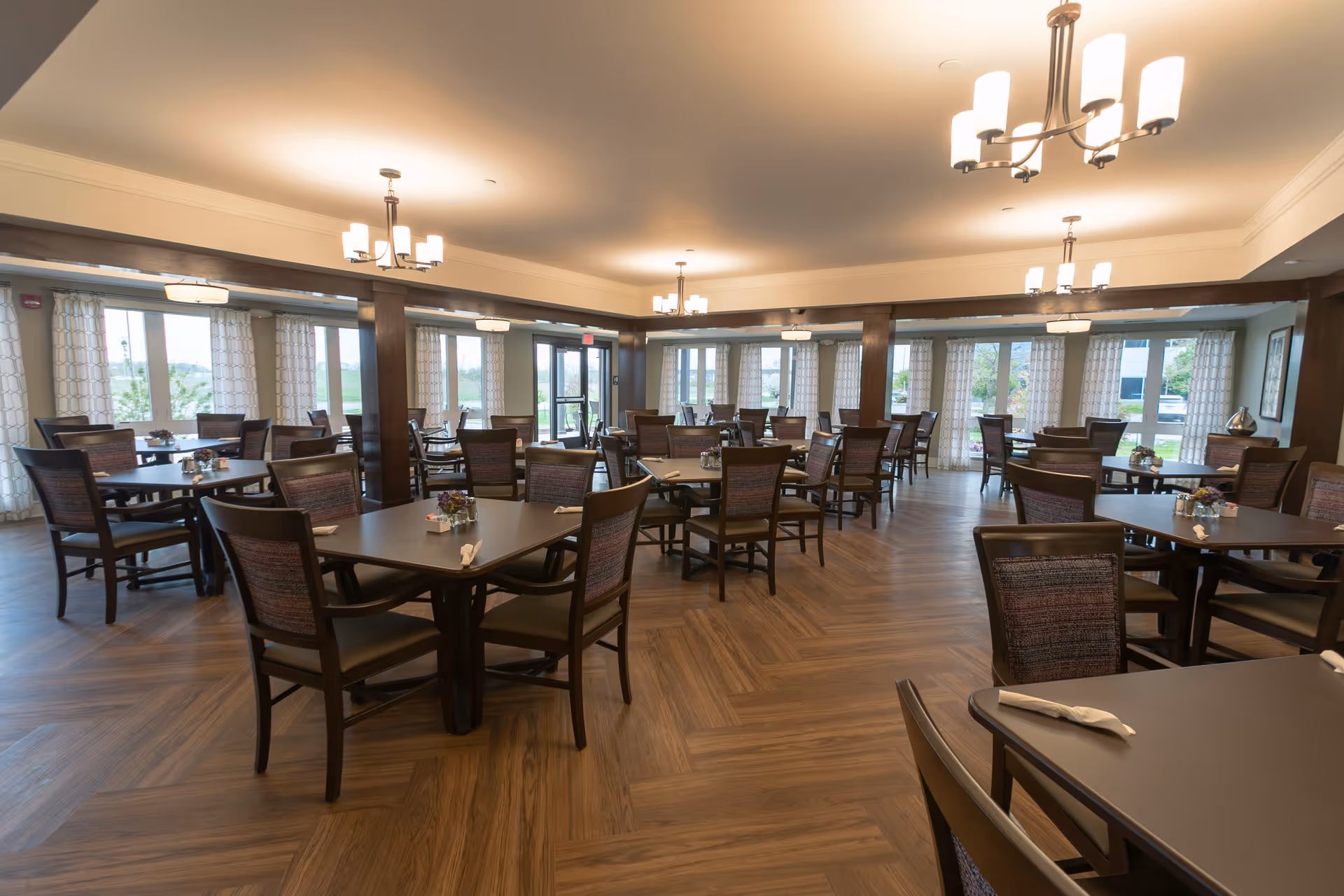A spacious dining room with multiple square tables and wooden chairs arranged neatly. The room features large windows with patterned curtains allowing natural light to fill the space. The floor has a wood-like finish, and modern ceiling lights hang from the ceiling providing warm illumination.