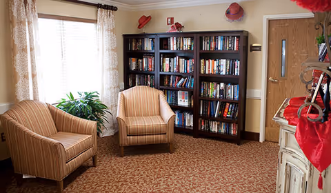 A cozy reading area with two striped armchairs, a bookcase filled with books, a window with curtains, and a potted plant.