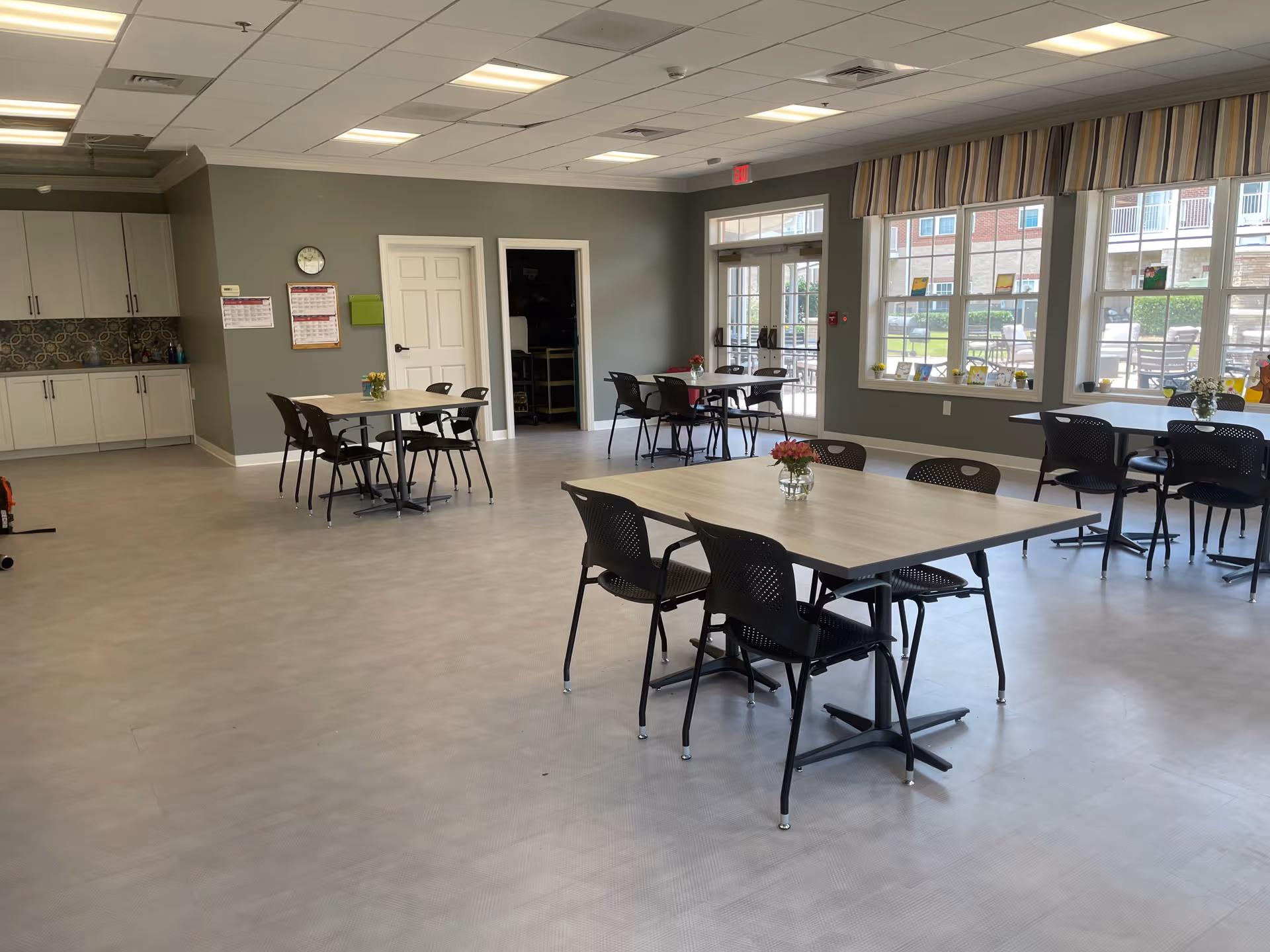 A bright dining room with several tables and black chairs arranged neatly. Each table has a small vase with flowers. Large windows on one side let in natural light and offer a view of an outdoor patio area. The walls are painted a muted green, and there is a door leading outside. Cabinets and a tiled backsplash are visible on the left side of the room.