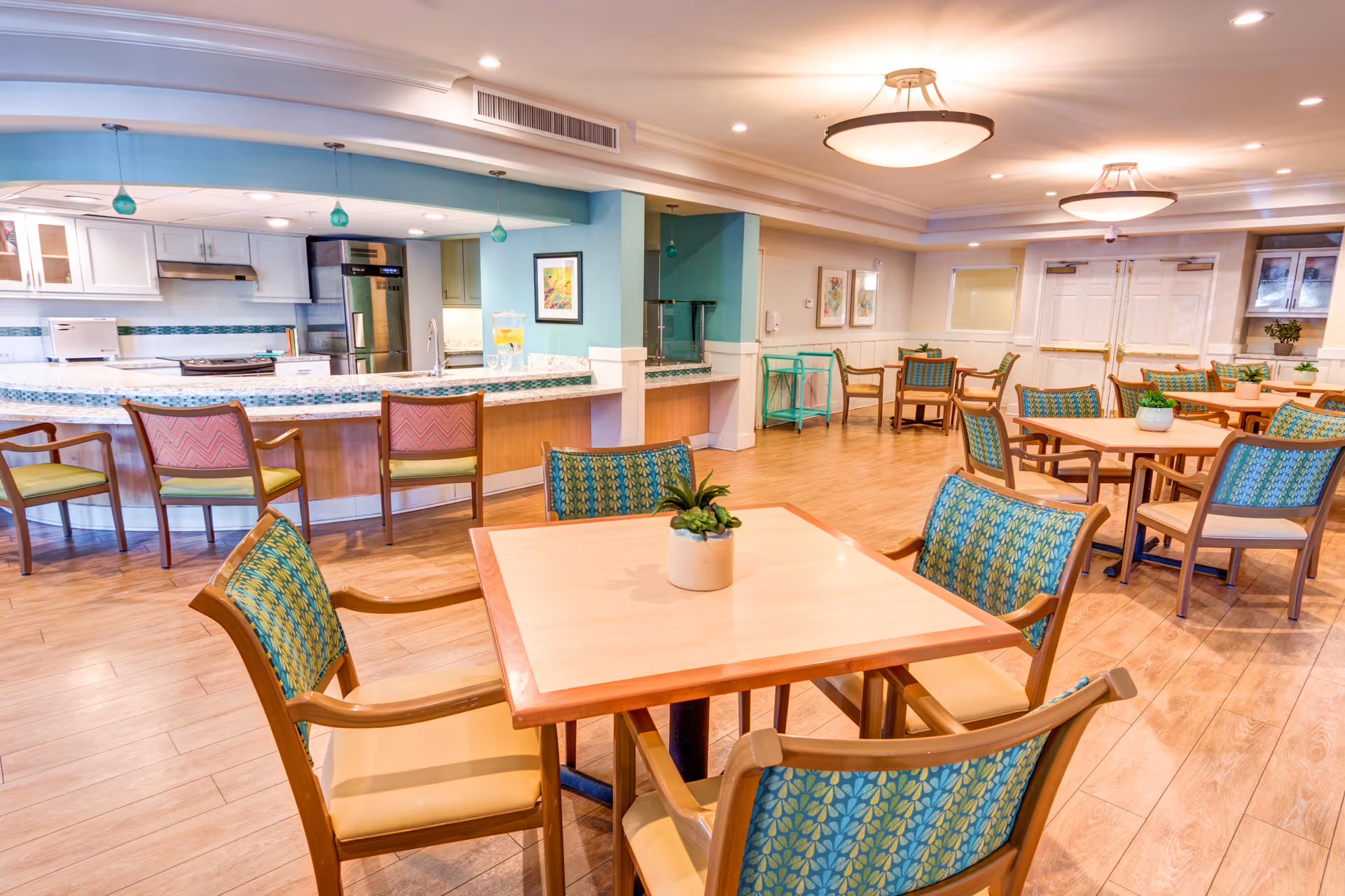 Bright communal dining area with multiple wooden tables and patterned chairs and an open kitchen/bar in the background.