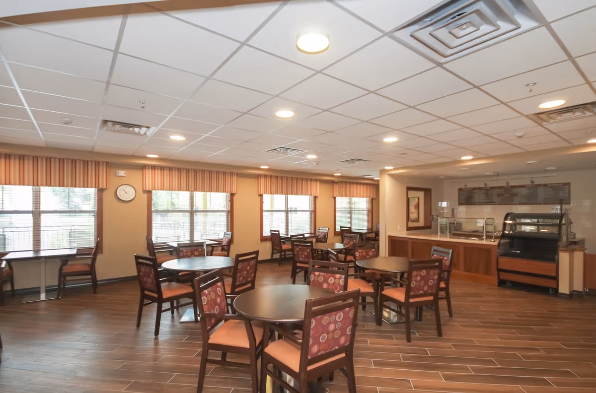 A dining room in a senior living facility with several round tables and chairs arranged neatly. The chairs have patterned upholstery with circular designs. Large windows with striped valances allow natural light to fill the room. There is a serving counter and a display case along one wall. The floor is covered with wood-like tiles, and the ceiling has recessed lighting.