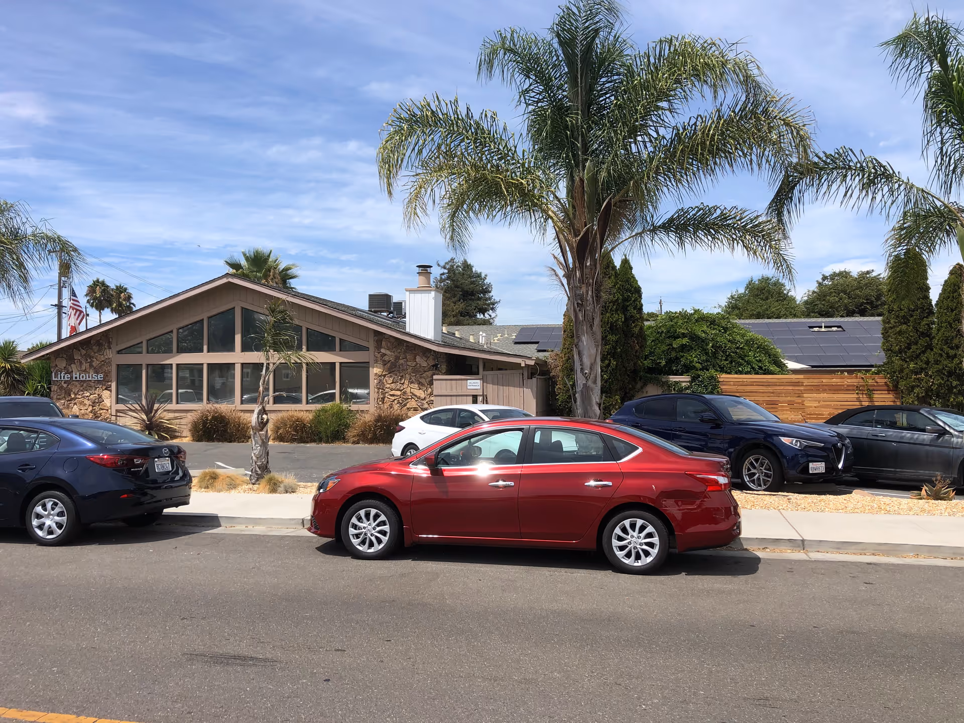 Exterior view of Solano Life House facility with a stone facade and large windows under a sloped roof. Several cars are parked along the street and in the parking area. Palm trees and other greenery surround the building under a partly cloudy sky.