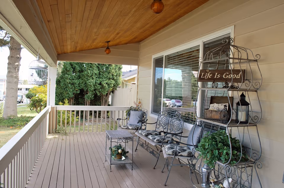 Covered front porch with metal chairs, a small table and a decorative shelf displaying a 'Life Is Good' sign.