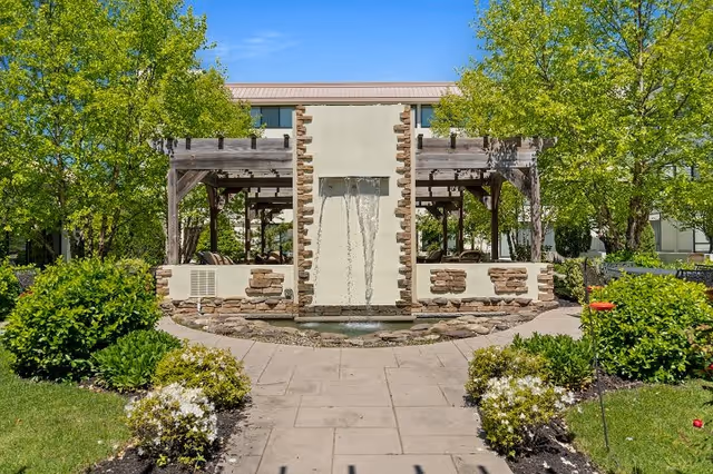Outdoor garden area with a central water feature consisting of a stone and stucco wall with water flowing from two spouts into a small pond. The area is surrounded by green bushes, trees, and a paved walkway leading to the water feature. There is a wooden pergola structure on either side of the water feature providing shaded seating areas.