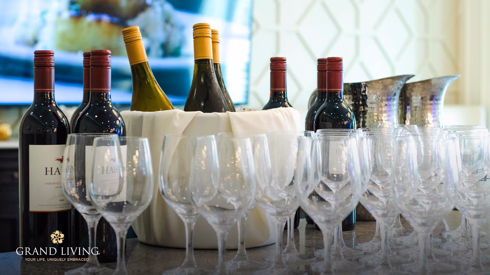 A collection of wine bottles and empty wine glasses arranged on a countertop, with some bottles placed in a white cloth-covered ice bucket. The background shows a blurred interior setting with a patterned wall.