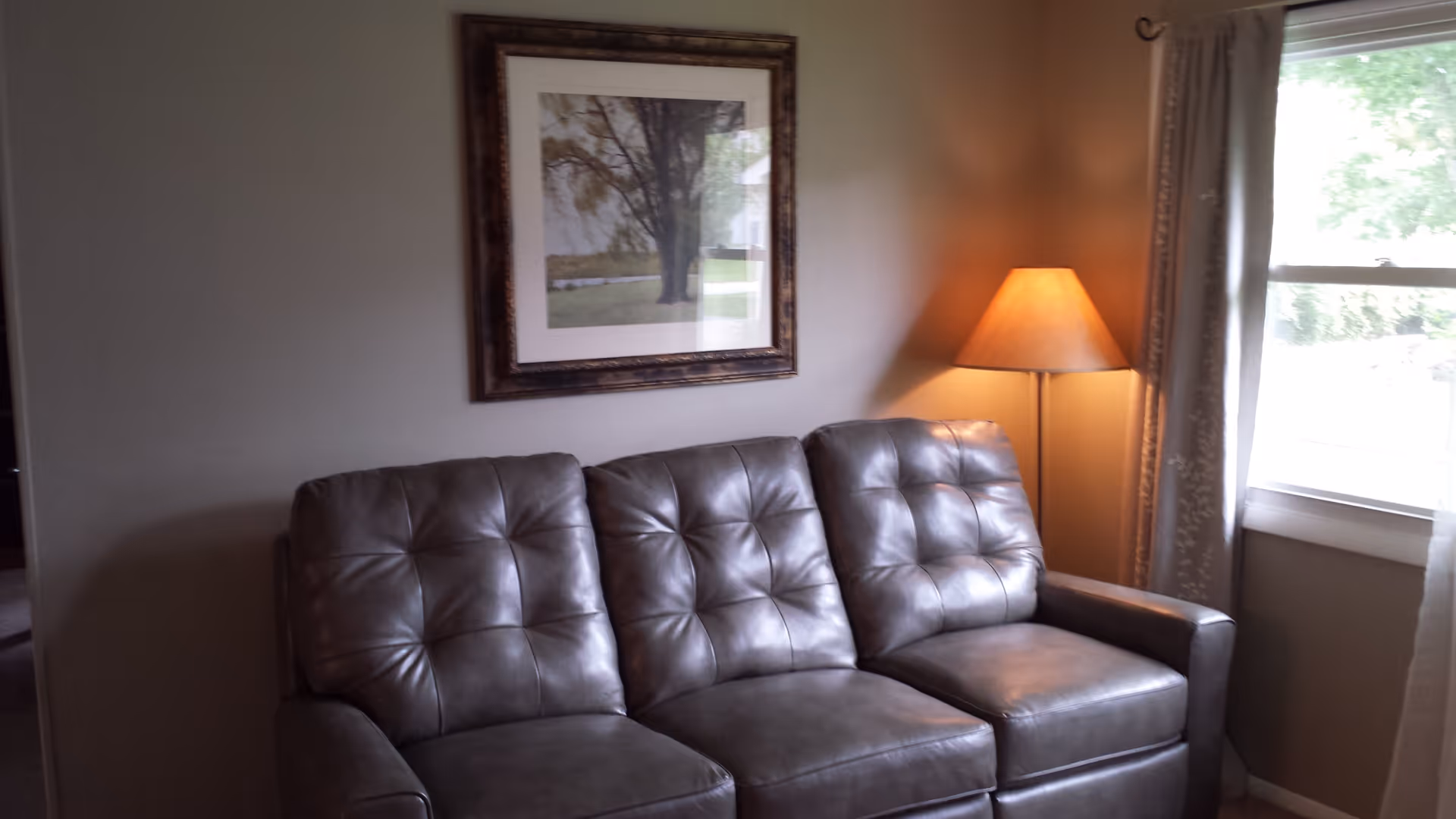 A cozy living room with a gray leather tufted sofa, a framed picture of a tree on the wall above the sofa, a lit floor lamp with a beige shade in the corner, and a window with curtains letting in natural light.