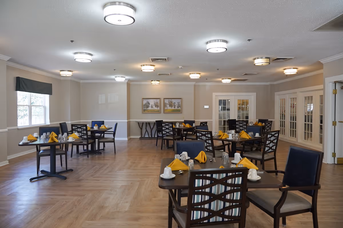 A spacious dining room with multiple dark wood tables and chairs arranged neatly. Each table is set with yellow folded napkins, white cups, glasses, and silverware. The room has light-colored walls, wood flooring, several ceiling lights, and windows with dark valances allowing natural light in. Two framed landscape paintings hang on the far wall.