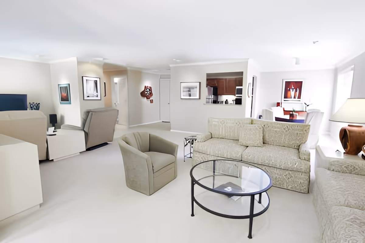 A bright and spacious living room in a senior living facility with beige sofas, a matching armchair, a glass coffee table, and a lamp on a side table. The room has light-colored walls and carpet, with framed artwork on the walls. In the background, there is a dining area with a table and chairs, and a kitchen visible through a pass-through window.